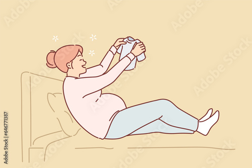Happy pregnant woman sits on bed holding small t-shirt for baby in hands and smiles