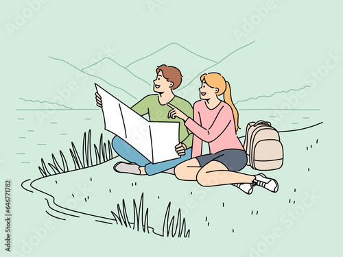 Happy couple of travelers sitting on lawn during hike in national park with lake and holding map in hands. Cheerful man and woman went on hike for adventure and new positive emotions