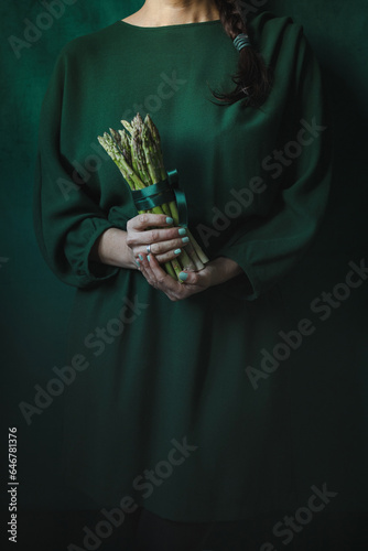 Woman in a green dress holding a bunch of fresh green asparagus with a dark green background