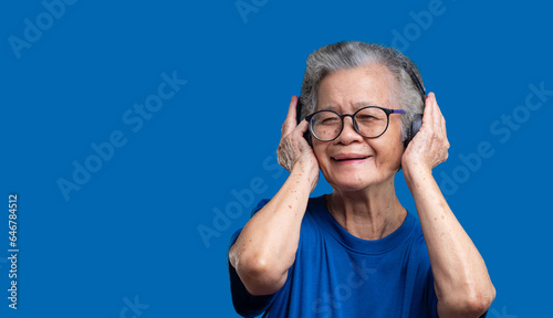 Cheerful senior woman wearing wireless headphones and hands up with a smile while standing on a blue background.