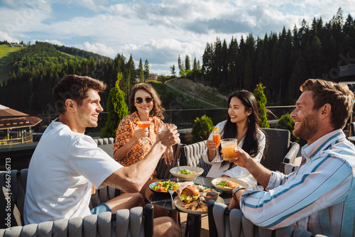 Group of friends drinking cocktails and enjoying meal while sitting on restaurant terrace in mountains
