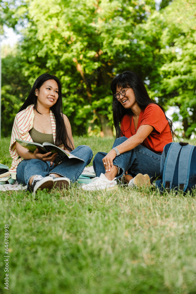 Two asian female students doing homework together while sitting on grass in park