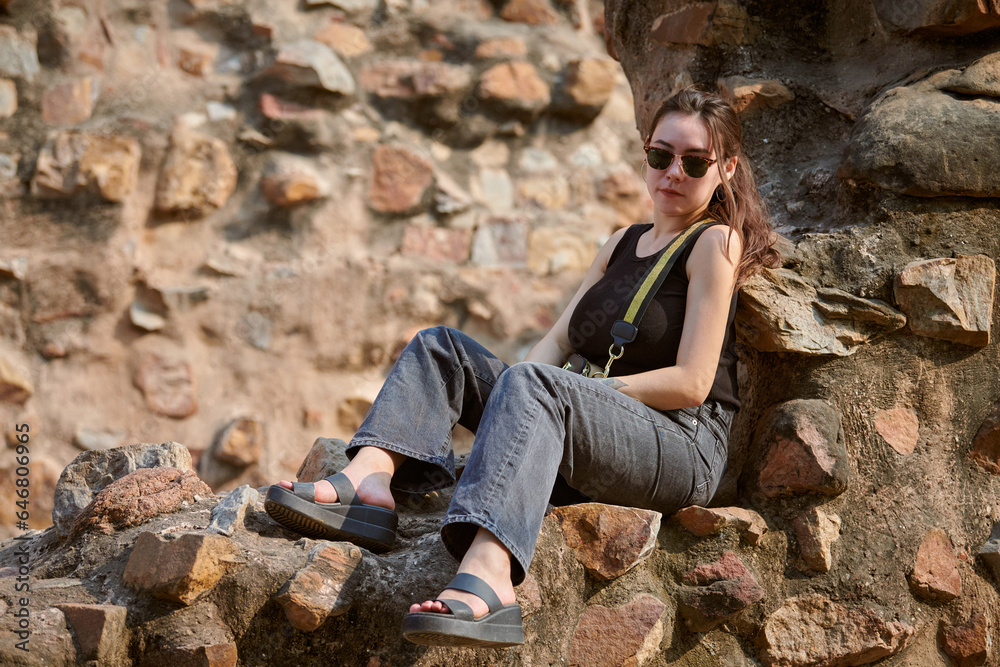 Attractive east asian woman in black sunglasses and black clothes sitting on ancient stone wall of old indian architecture buildings in Qutb Minar complex, candid young girl photo session in India Attractive east asian woman in black sunglasses and black clothes sitting on ancient stone wall of old indian architecture buildings in Qutb Minar complex, candid young girl photo session in India