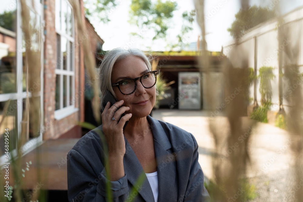 charming slim mature woman with glasses dressed in a gray jacket and jeans holding a smartphone outdoors