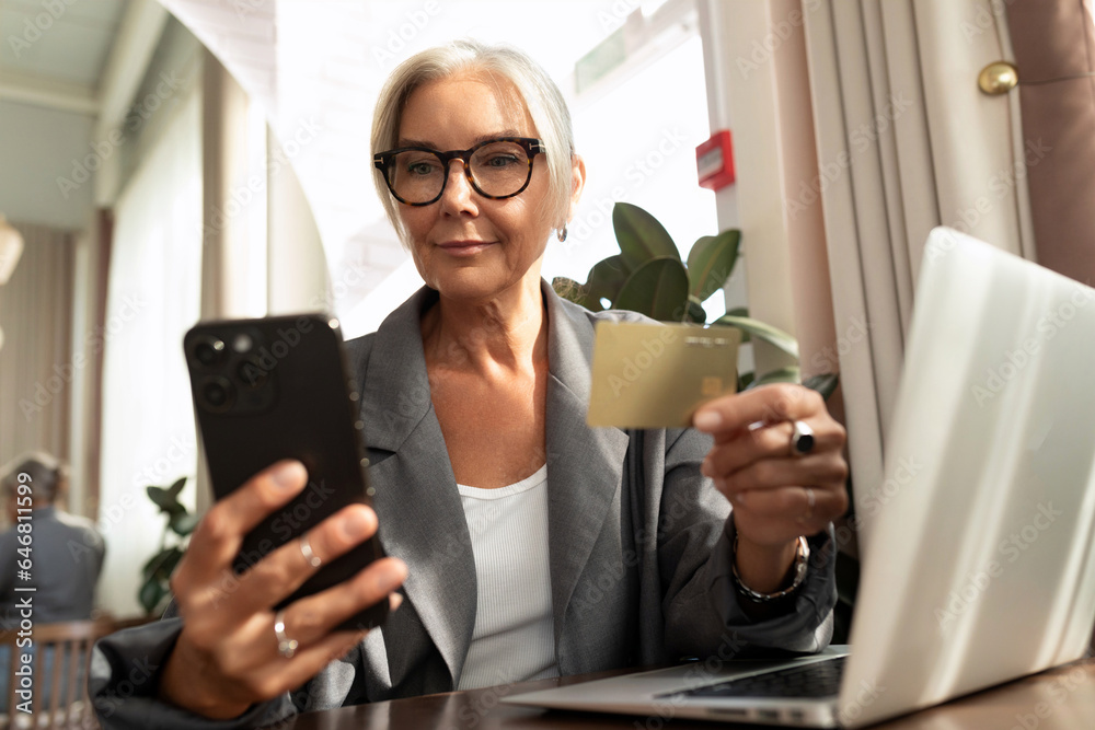 gray-haired mature pretty business woman dressed in a stylish gray jacket pays the bill in a cafe using a smartphone and a plastic card gray-haired mature pretty business woman dressed in a stylish gray jacket pays the bill in a cafe using a smartphone and a plastic card