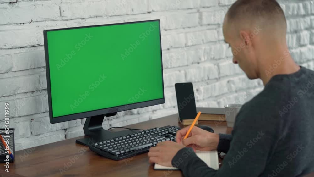 Man Looks at Computer Green Screen and Writes Something in Notebook. Young Man Works In Modern Office With White Brick Wall Man Looks at Computer Green Screen and Writes Something in Notebook. Young Man Works In Modern Office With White Brick Wall