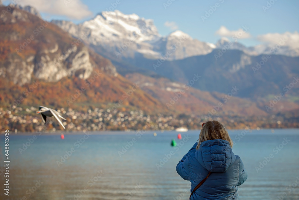 A senior woman stands by Lake Annecy, capturing the mountainous landscape on her mobile phone. Elder traveler in the serene beauty of the French Alps