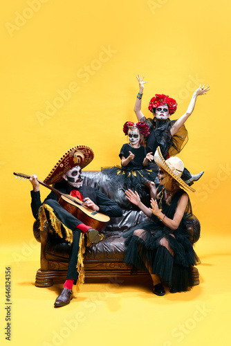 Skeletons, zombies family, parents and children sitting on sofa singig and dancing with guitar celebrating Halloween over bright yellow background.
