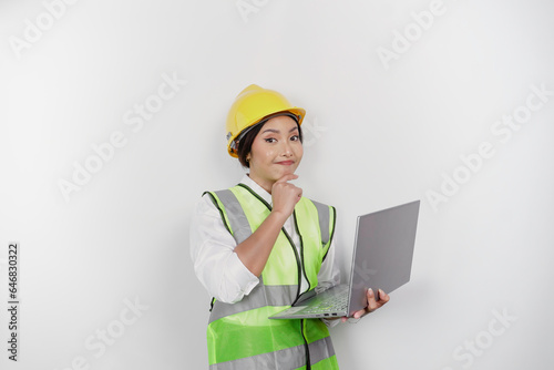 A thoughtful young woman labor worker wearing safety helmet and vest while holding her laptop and hand on her chin  isolated by white background. Labor s day concept.
