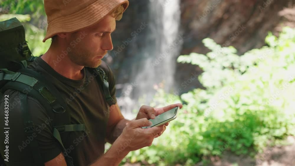 Concentrated tourist blond man in panama hat looking at route on map on phone against waterfall in mountains Concentrated tourist blond man in panama hat looking at route on map on phone against waterfall in mountains