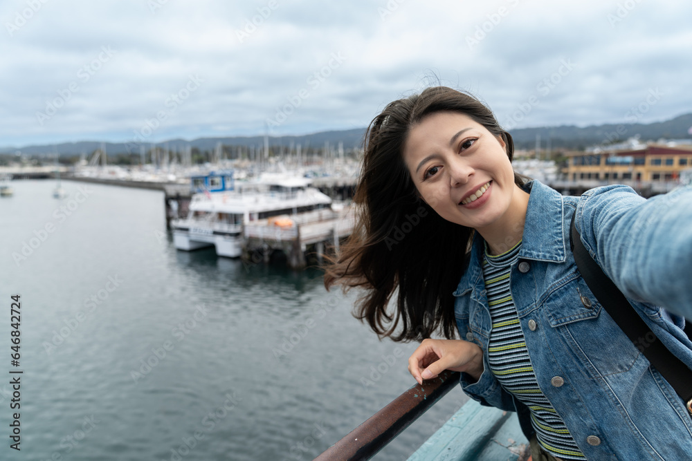 self portrait of happy asian woman visitor smiling at camera while taking selfie picture with boats near fishing port at Old Fisherman's Wharf in Monterey California usa