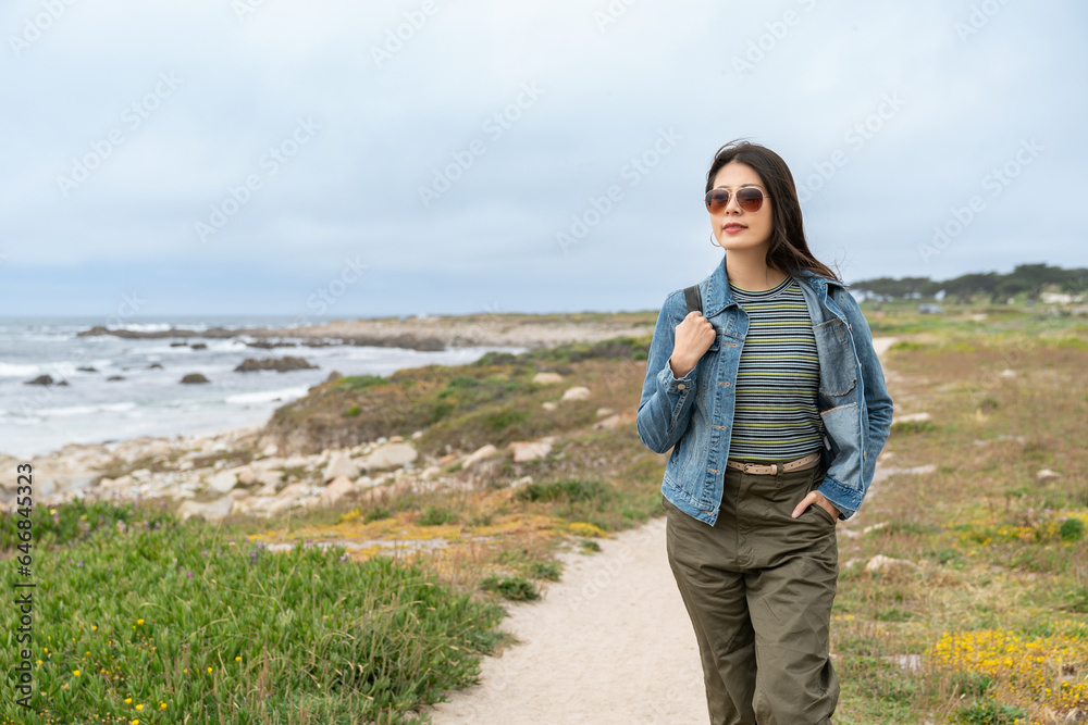 fashionable asian Japanese girl traveler walking leisurely along scenic California coastal beach covered in grass in united states with hand in pocket on a breezy day fashionable asian Japanese girl traveler walking leisurely along scenic California coastal beach covered in grass in united states with hand in pocket on a breezy day