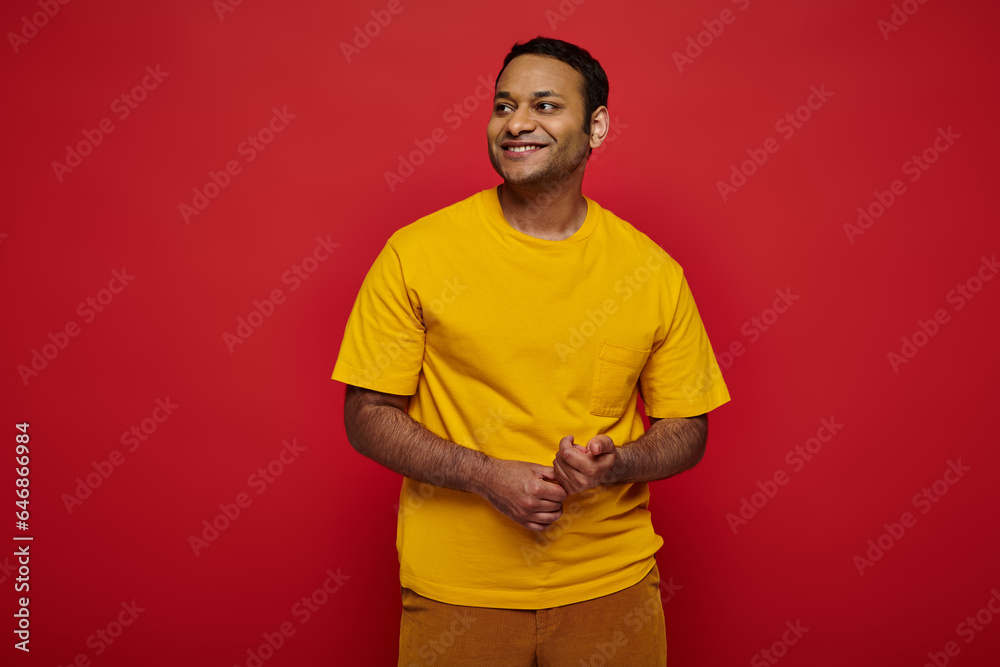 delightful indian man in bright casual clothes looking away and smiling on red background in studio