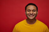 positive emotion, happy indian man in yellow t-shirt smiling or grinning on red background in studio