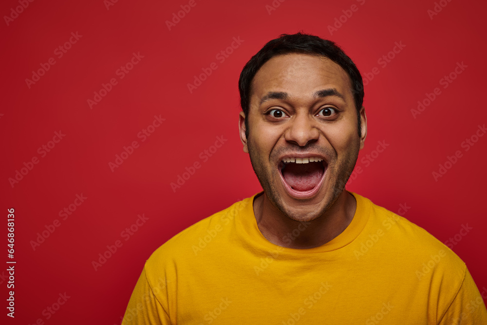 positive emotion, excited indian man in yellow t-shirt with opened mouth on red background