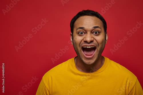 positive emotion, excited indian man in yellow t-shirt with opened mouth on red background