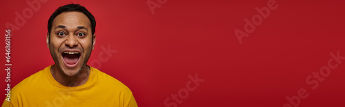 positive emotion, excited indian man in yellow t-shirt with opened mouth on red background, banner