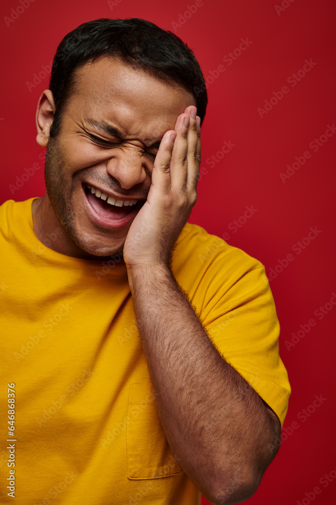 face expression, embarrassed indian man in yellow t-shirt laughing and touching face on red backdrop face expression, embarrassed indian man in yellow t-shirt laughing and touching face on red backdrop
