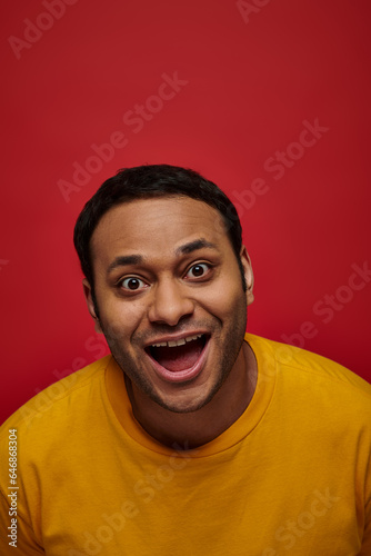 positive emotion, excited indian man in yellow t-shirt with opened mouth on red background, vertical