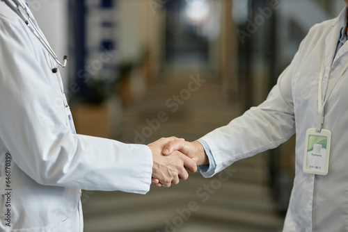 Close-up of hands of two general practitioners in lab coats standing in front of camera in corridor of modern hospital during handshake