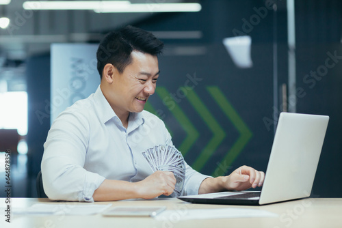 Happy young Asian businessman working in office on laptop and holding cash money banknotes in hands.