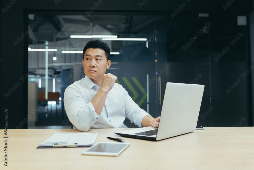 Portrait of a young Asian male businessman in a white shirt working in the office at a laptop. He looks seriously to the side. Portrait of a young Asian male businessman in a white shirt working in the office at a laptop. He looks seriously to the side.