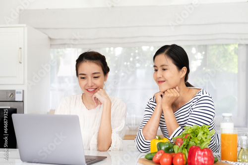 Ordering Food Online. Smiling girlfriends with laptop purchasing grocery delivery from internet while cooking together in kitchen