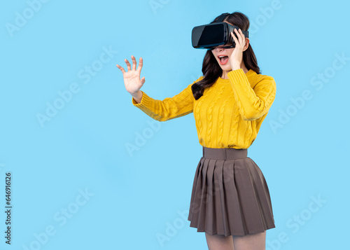 Excited young woman in VR headset touching air during virtual reality experience at studio. Emotional lady exploring artificial computer world or playing video game isolated on light blue background