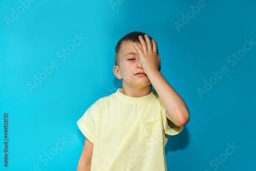 A 6-7-year-old boy in a yellow T-shirt shows a gesture of being bored covering his face with one hand on a blue background. Close-up portrait, front view