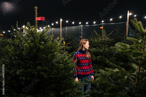 A young woman in a sweater looks at the Christmas trees in the middle of the Christmas market against the background of garlands. Winter wonder holiday concept photo