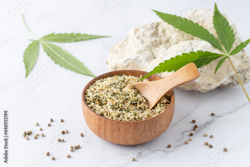 Shelled crushed hemp seeds in wooden bowl as ingredient for eating and cannabis leaves on white marble background