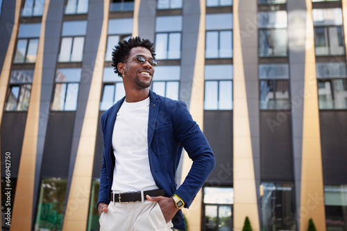 Confident african American male in stylish clothes stands in city next to modern building, looking to side with smile. Happy black man with hands in pockets