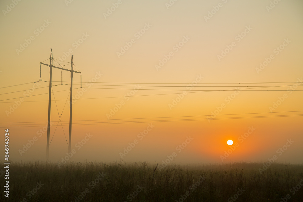 The setting sun in the field against the background of power lines evening dark golden sky and fog.
