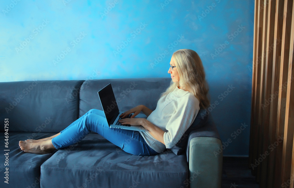 Modern middle-aged gray-haired woman in eyeglasses working with laptop sitting on the couch in the room at home Modern middle-aged gray-haired woman in eyeglasses working with laptop sitting on the couch in the room at home