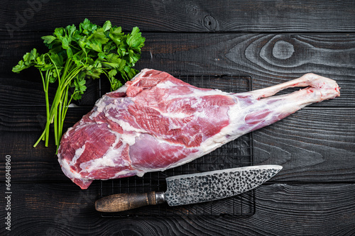 Fresh Raw whole lamb mutton leg thigh on kitchen table with herbs. Black Wooden background. Top view