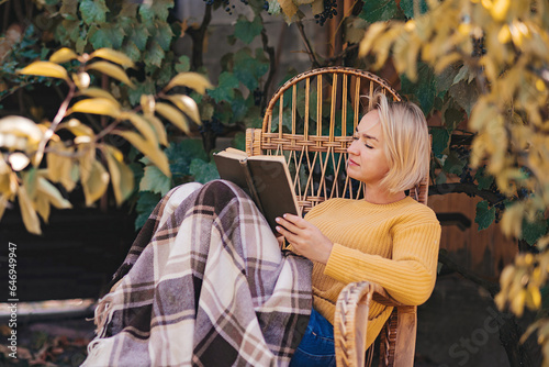 Beautiful girl resting sitting in autumn garden in chair wrapped in a woolen plaid blanket. Girl reading a book