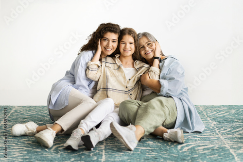 Glad caucasian diverse women hugs, enjoy love and support, sit on floor on white wall background, full length