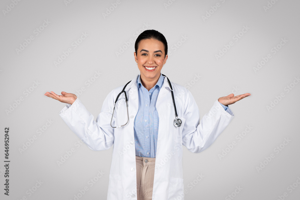 Friendly brazilian female doctor in uniform showing both hands open palms, leaving choice to patient, grey background Friendly brazilian female doctor in uniform showing both hands open palms, leaving choice to patient, grey background