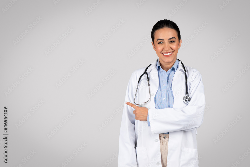 Useful information. Portrait of cheerful latin female doctor pointing at copy space, grey background, smiling at camera Useful information. Portrait of cheerful latin female doctor pointing at copy space, grey background, smiling at camera