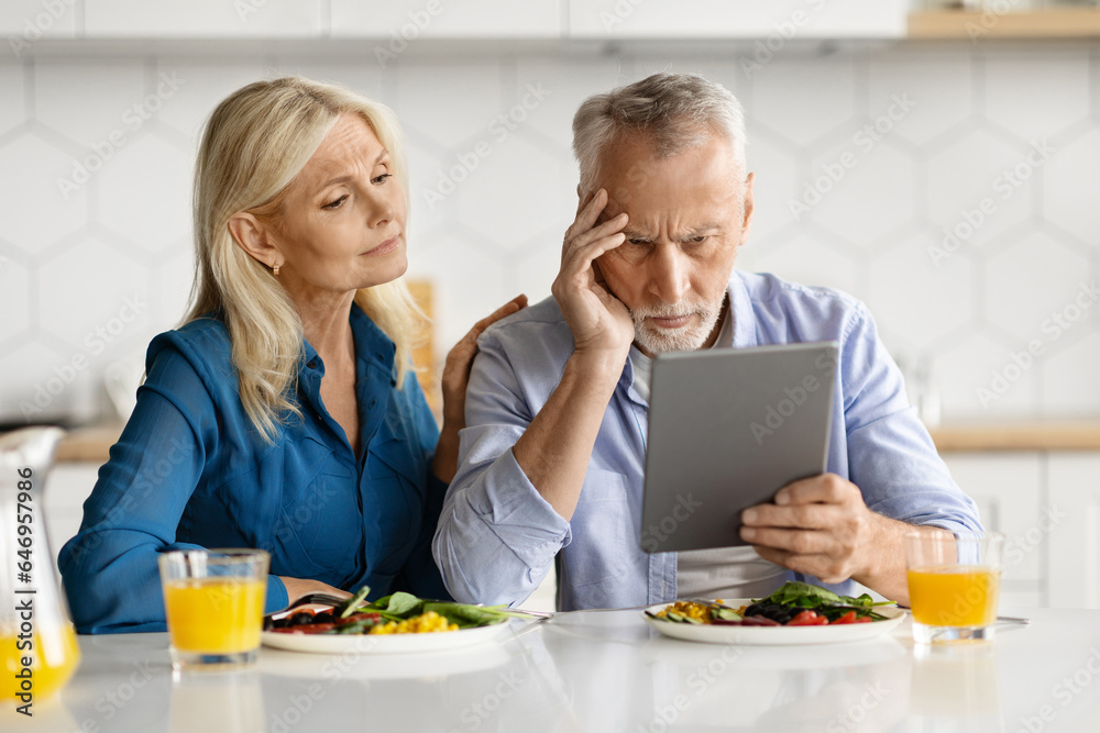 Upset senior spouses using digital tablet while eating breakfast in kitchen