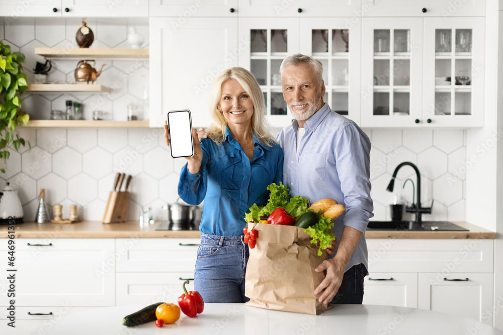 Smiling Senior Spouses Recommending Grocery Delivery App, Showing Blank Smartphone In Kitchen Smiling Senior Spouses Recommending Grocery Delivery App, Showing Blank Smartphone In Kitchen