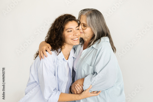Positive caucasian old woman kiss in cheek millennial lady, enjoy love and support on white wall background
