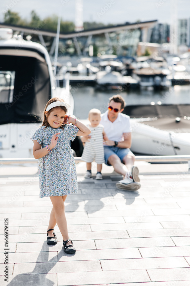 A child girl is walking with her dad and little brother on the street. A beautiful little girl is wearing a summer dress. Happy child resting with his father and brother A child girl is walking with her dad and little brother on the street. A beautiful little girl is wearing a summer dress. Happy child resting with his father and brother
