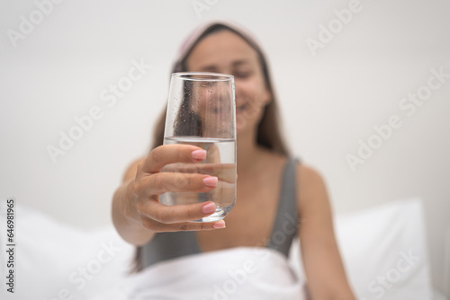 Smiling young woman holds out glass of water sitting on bed with light sleeping linen in bedroom of accommodation taking water after receiving energy from sleep