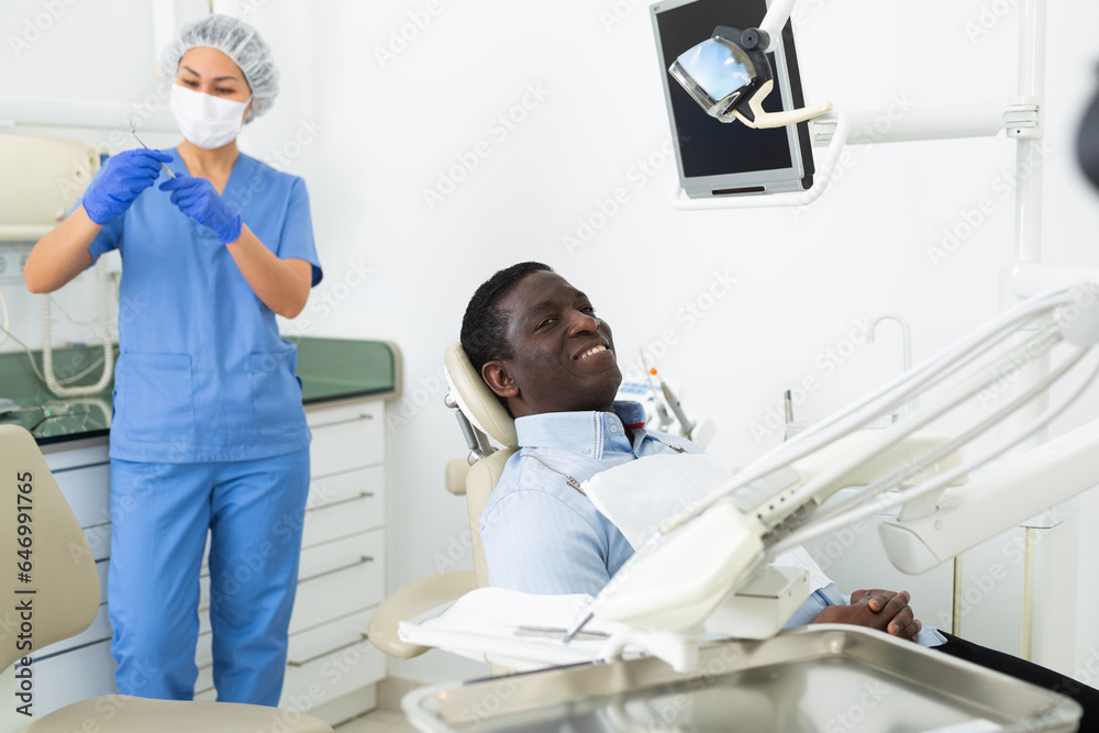 Fototapeta premium Positive african-american man patient sitting on dental chair and smiling while Asian woman dentist standing in background and preparing tools for examination.
