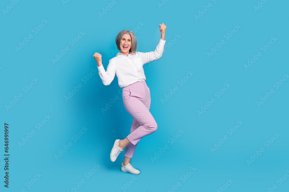 Full body portrait of delighted aged lady raise fists achieve luck empty space isolated on blue color background Full body portrait of delighted aged lady raise fists achieve luck empty space isolated on blue color background
