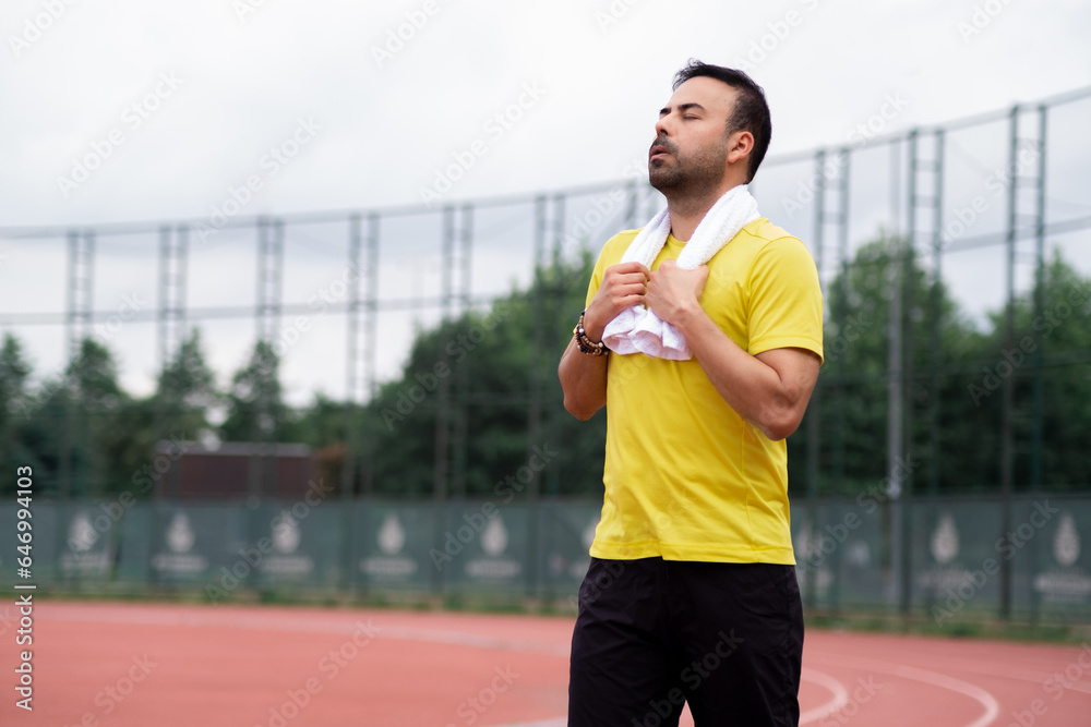 Tired sportsman with white towel around neck resting walking along running track after training at city stadium professional athlete doing everyday workout on outdoor arena Tired sportsman with white towel around neck resting walking along running track after training at city stadium professional athlete doing everyday workout on outdoor arena