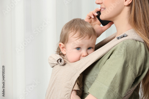 Mother talking on smartphone while holding her child in sling (baby carrier) indoors
