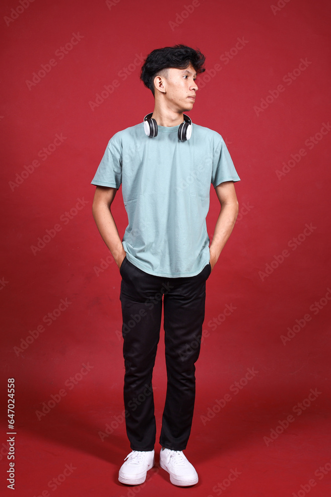 Fototapeta premium Full length of smiling young Asian man wearing headphones looking aside isolated on crimson red background
