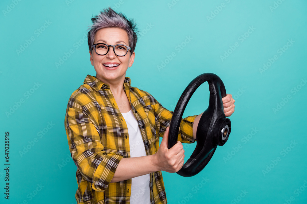 Photo of lovely senior lady hold steering wheel rudder dressed stylish checkered yellow garment isolated on aquamarine color background Photo of lovely senior lady hold steering wheel rudder dressed stylish checkered yellow garment isolated on aquamarine color background